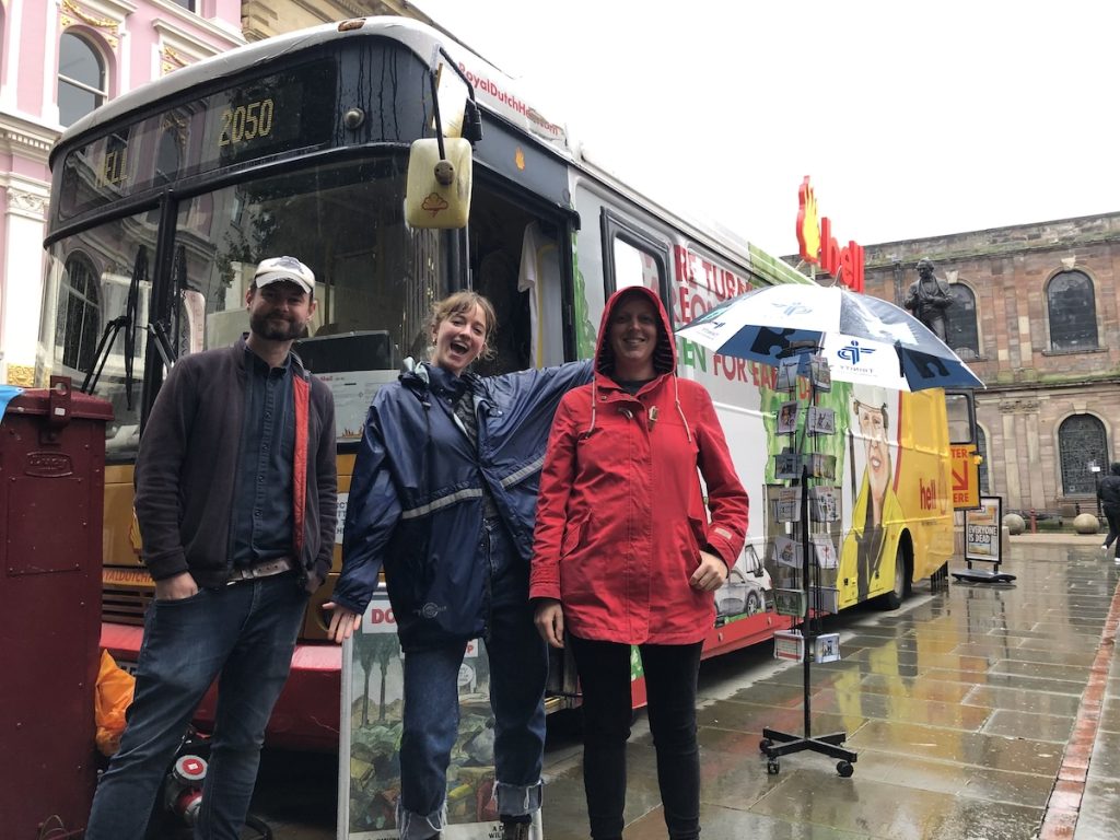 Three people stand in front of a yellow bus that says "Hell"